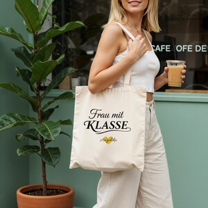 Può includere: Borsa tote color crema con il testo tedesco "Frau mit Klasse" in corsivo nero, un cuore dorato e il nome "Angelika". La borsa è portata da una persona che indossa un top e pantaloni bianchi. Una pianta in vaso di terracotta è sullo sfondo.