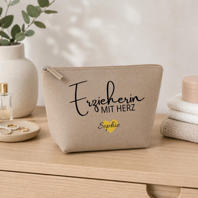 May include: A beige cosmetic bag with a zip closure. The bag displays the text "Erzieherin MIT HERZ" in black script, with a yellow heart and the name "Sophie" below. The bag is on a wooden surface.