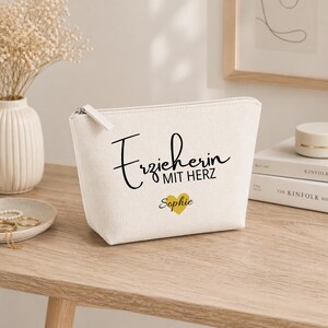 May include: A cream-coloured zippered cosmetic bag with the text "Erzieherin MIT HERZ" in black script, a gold heart, and the name "Sophie". The bag is on a wooden surface.