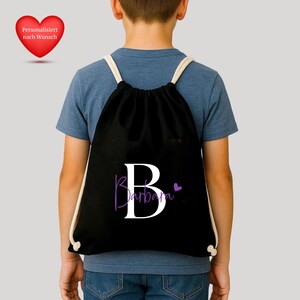 Op de afbeelding: Zwarte rugzak met trekkoord met een grote witte letter "B" en de naam "Barbara" in paars schrift. Een klein paars hartje staat rechts van de naam. De rugzak heeft witte trekkoorden.