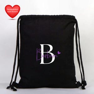 Op de afbeelding: Zwarte rugzak met trekkoord met een grote witte letter "B" en de naam "Barbara" in paars schrift. Een klein paars hart staat naast de naam. Een rood hart met Duitse tekst staat in de linkerbovenhoek.