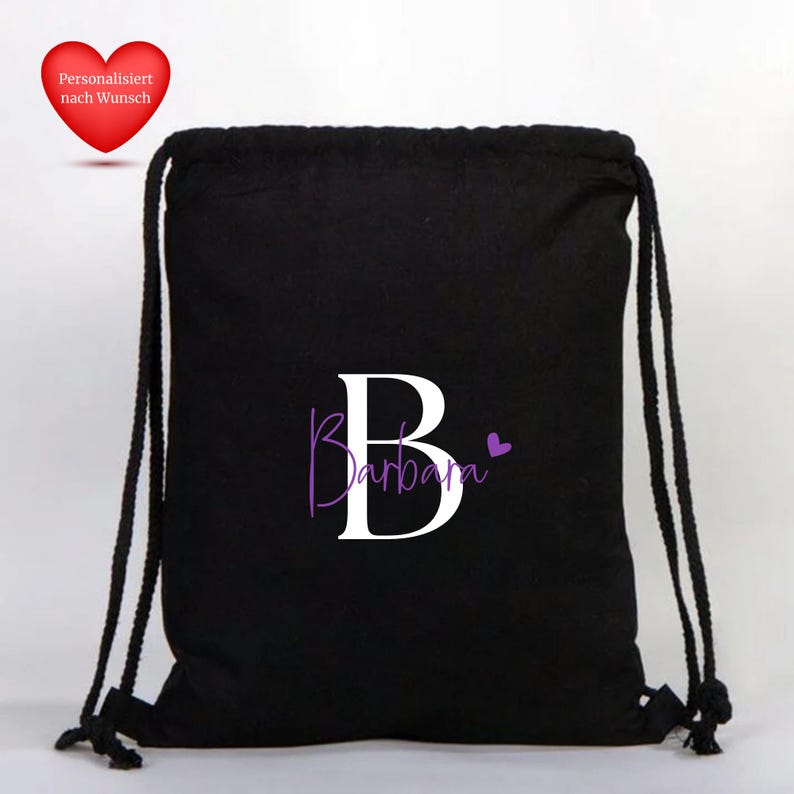 Op de afbeelding: Zwarte rugzak met trekkoord met een grote witte letter "B" en de naam "Barbara" in paars schrift. Een klein paars hart staat naast de naam. Een rood hart met Duitse tekst staat in de linkerbovenhoek.