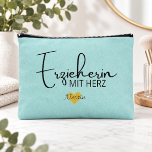 May include: A light blue rectangular zippered pouch featuring the black text "Erzieherin MIT HERZ" and a yellow heart icon above the name "Nesrin" in black script. The pouch has a black zipper closure along the top edge and a textured fabric surface.