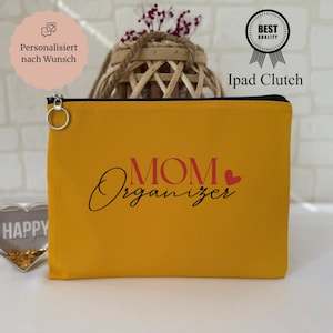 Può includere: Pochette per iPad giallo senape con cerniera nera e anello argentato. Il testo "MOM Organizer" è stampato in rosso e nero. Un cuore rosso è accanto alla parola "Organizer". Il testo "iPad Clutch" è stampato in nero.