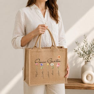 Pode incluir: Uma bolsa de juta bege com o texto preto Omas Garten acima de cinco ilustrações de flores coloridas em estilo aquarela. Cada caule de flor está etiquetado com um nome em letra cursiva: Miguel, Jonathan, Linnea, Bruno e Lucas.