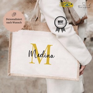 Könnte beinhalten: Eine personalisierte Tragetasche mit dem Namen "Medina" in schwarzer Schrift und einem goldenen "M". Die Tasche hat eine natürliche Jute-Verzierung und Griffe, mit einer weißen Leinwandfront. Das Bild enthält auch ein "Best Quality"-Siegel.