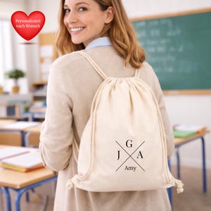 Puede incluir: Una mochila con cordón de color crema con las iniciales G, J y A, y el nombre Amy impreso en negro. La mochila la lleva una persona en un aula. El texto "Personalisiert nach Wunsch" está en un corazón rojo.