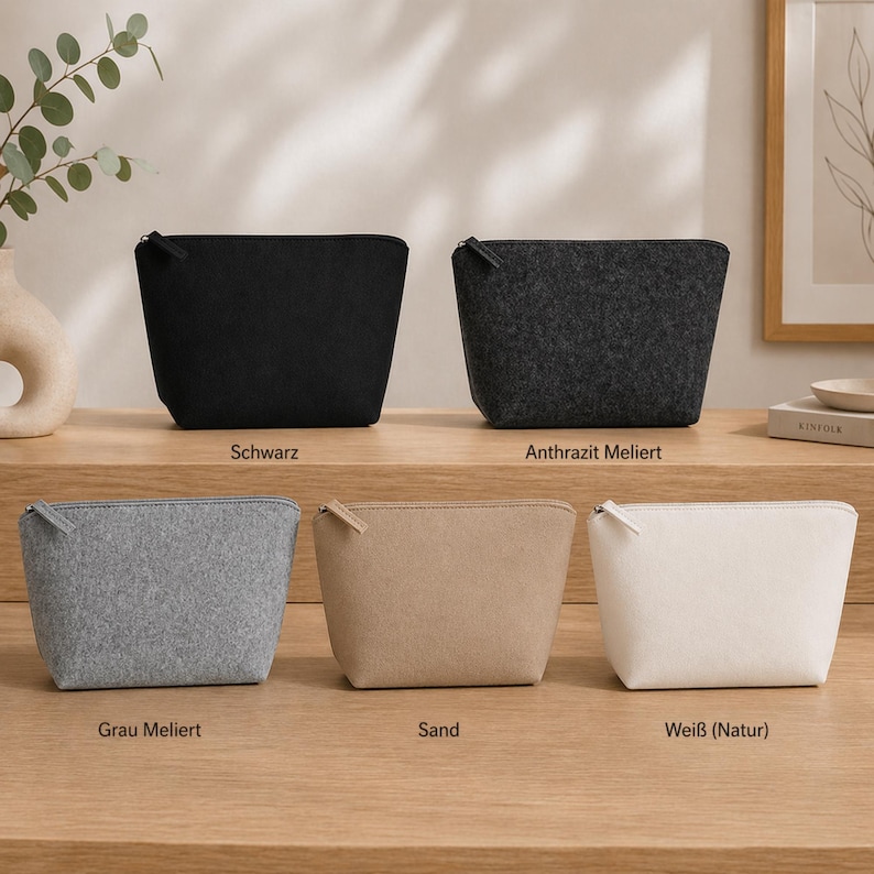 May include: Five felt cosmetic bags in black, dark grey, light grey, tan, and white. Each bag has a zip closure and is displayed on a wooden surface. The bags are labelled with their respective colour names.