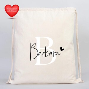 Op de afbeelding: Een cr&egrave;mekleurige trekkoordtas met de naam "Barbara" in een zwart scriptlettertype, over de grote witte letter "B" geplaatst. Een klein zwart hartje staat naast de naam. Een rood hart met tekst staat linksboven.