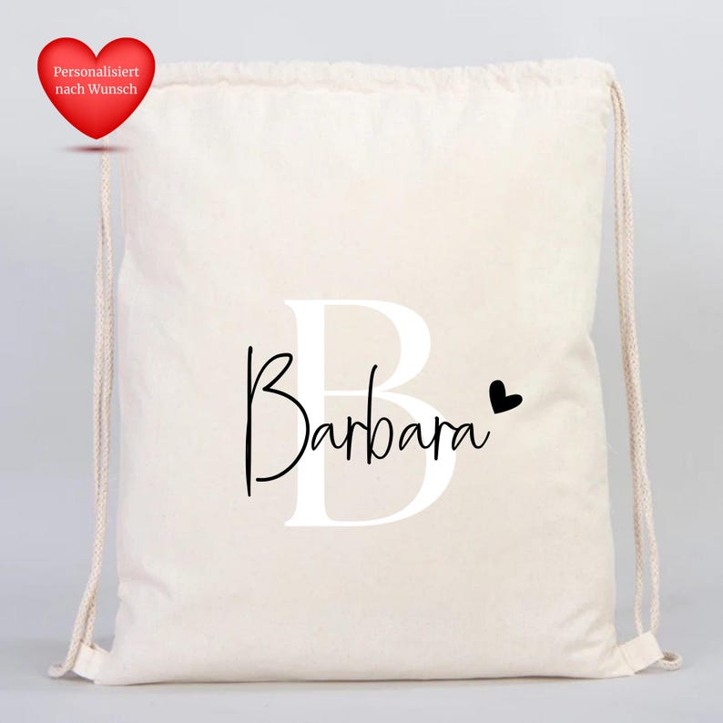 Op de afbeelding: Een cr&egrave;mekleurige trekkoordtas met de naam "Barbara" in een zwart scriptlettertype, over de grote witte letter "B" geplaatst. Een klein zwart hartje staat naast de naam. Een rood hart met tekst staat linksboven.