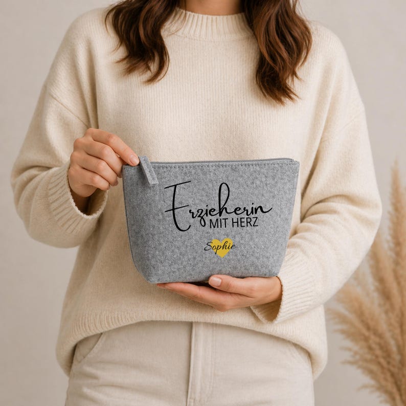 Könnte beinhalten: Eine graue Filz-Reißverschlusstasche mit dem Text "Erzieherin mit Herz" und dem Namen "Sophie" unter einem gelben Herz. Die Tasche wird von einer Person gehalten, die einen cremefarbenen Pullover trägt.