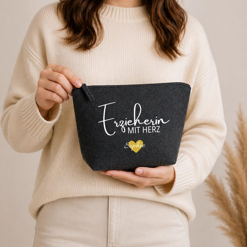 May include: A dark grey felt cosmetic bag with a zip closure. The bag features white script text that reads "Erzieherin MIT HERZ" and a gold heart with the name "Sophie" below. The bag is held by a person wearing a cream-coloured sweater.