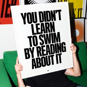 Może przedstawiać: Prostokątny biały plakat z pogrubionym czarnym tekstem o treści: YOU DIDN'T LEARN TO SWIM BY READING ABOUT IT. Plakat jest trzymany pionowo, prezentując czysty, kontrastowy projekt z prostym krojem pisma bezszeryfowego na białym tle.
