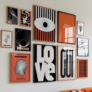 Op de afbeelding: Een galerijmuur met ingelijste kunstprints met verschillende ontwerpen en tekst, waaronder "LOVE", "FOCUS" en "OVERTHINKER". Het kleurenpalet omvat zwart, wit en oranje. Een oranje bank met patronen kussens staat eronder.