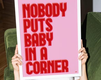 May include: A rectangular wall art print in a white frame featuring a light pink background with the bold red text NOBODY PUTS BABY IN A CORNER. The lettering has a slight drop shadow effect, creating a layered appearance on the flat surface.