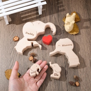 K&ouml;nnte beinhalten: Holzpuzzle einer Elefantenfamilie mit den Namen "Daddy", "Mummy", "Lucas" und "Ella", die auf jedes Teil graviert sind. Ein rotes Herz ist im Puzzle enthalten.