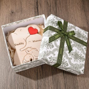K&ouml;nnte beinhalten: Ein Holzpuzzle in Form einer Elefantenfamilie in einer wei&szlig;en Geschenkbox mit gr&uuml;nem Band. Die Puzzleteile sind mit "Papa", "Mama" und "Lucas" beschriftet.