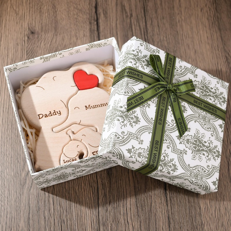 K&ouml;nnte beinhalten: Ein Holzpuzzle in Form einer Elefantenfamilie in einer wei&szlig;en Geschenkbox mit gr&uuml;nem Band. Die Puzzleteile sind mit "Papa", "Mama" und "Lucas" beschriftet.