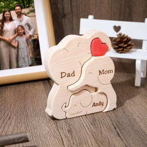 K&ouml;nnte beinhalten: Holzpuzzle einer Elefantenfamilie mit den W&ouml;rtern "Papa", "Mama" und "Andy", die auf jeden Elefanten graviert sind. Die Elefanten halten ein rotes Herz.