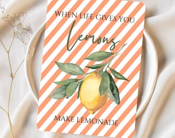 May include: A motivational quote card with a peach and white striped background. The card features a watercolor illustration of a lemon branch with green leaves and a yellow lemon. The text on the card reads "When life gives you lemons, make lemonade."