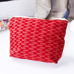May include: A rectangular zipped pouch made of red fabric featuring a repeating pattern of small, light-coloured arrow-like shapes. The pouch has a white zip closure along the top edge and a soft, structured form suitable for holding small items.