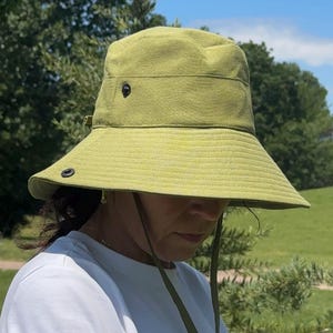 Könnte beinhalten: Ein hellgrüner Fischerhut mit breiter Krempe und Kinnriemen. Der Hut hat ein schlichtes Design mit einem dunklen Knopf an der Seite. Die Person trägt ein weißes T-Shirt. Der Hintergrund ist ein Park mit Bäumen und blauem Himmel.