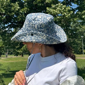 May include: A blue and white floral print sun hat with a wide brim. The hat has a chin strap and is worn outdoors. The hat is a stylish accessory for sunny days and outdoor activities.