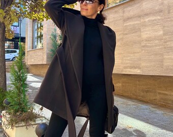 May include: A dark brown coat with a wide lapel and a matching belt. The coat is worn over a black turtleneck and black leggings. The outfit is completed with black knee-high boots and sunglasses. The background features a tree with yellow leaves.