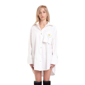 Op de afbeelding: Een vrouw draagt een wit oversized shirt met een bloemenontwerp op de zak en zwarte laarzen tot aan de knie.
