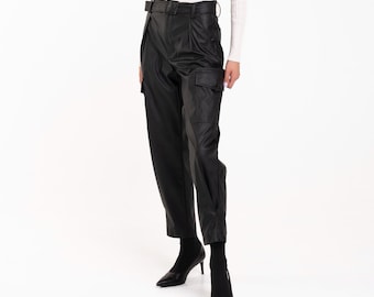 May include: A woman wearing a white turtleneck sweater and black leather pants with a belt. She is wearing black heels and has blonde hair.