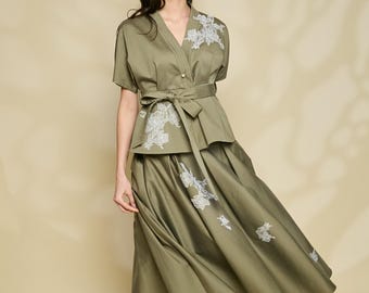 以下が含まれることがあります： オリーブグリーンのツーピースの衣装。ショートスリーブのラップトップスと、お揃いのミディスカートが特徴です。トップスとスカートには白い花の刺繍が施されています。お揃いのベルトでウエストを締めています。