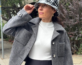 May include: A woman wearing a gray wool jacket with large pockets and button closures, paired with a black pleated skirt and a white turtleneck. She is also wearing a black and white patterned bucket hat.