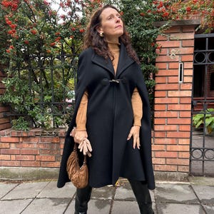 May include: A black cape coat with a clasp closure. The coat is worn over a tan turtleneck and paired with black boots. A brown handbag is held in the hand.