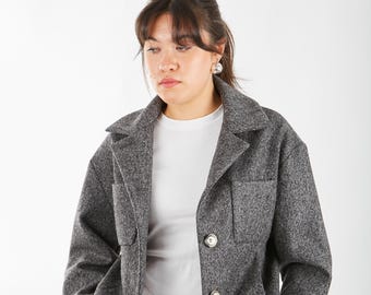 May include: A gray, cropped,  wool jacket with a button closure and a patch pocket on the left side. The jacket is worn over a white t-shirt and blue jeans.