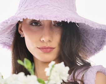 May include: A woman wearing a light purple, textured bucket hat with a frayed edge. She is holding a bouquet of white flowers.