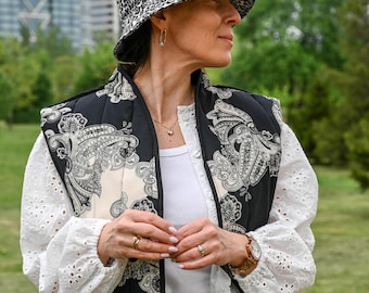 May include: A woman wearing a white blouse with eyelet detailing, a black and white paisley print quilted vest, blue jeans, and a black and white leopard print bucket hat.