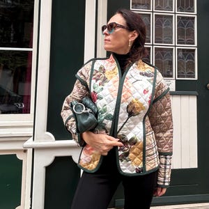 Puede incluir: Chaqueta acolchada con estampado floral en tonos rosa, amarillo y verde. Las mangas presentan un estampado de cuadros marrones y la chaqueta tiene un ribete verde. Se sostiene un pequeño bolso de mano verde oscuro.