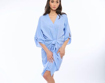 Puede incluir: Una mujer con un vestido azul claro hasta la rodilla con un lazo en la cintura. Está de pie sobre un fondo blanco y lleva sandalias blancas.