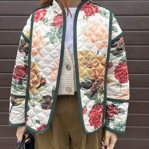 Op de afbeelding: Gewatteerde jas met een bloemenpatroon van rozen in rode, gele en perziktinten op een witte achtergrond. De jas heeft een donkergroene bies en een ruitvormig stiksel. De jas is open en onthult een wit shirt en een beige vest.