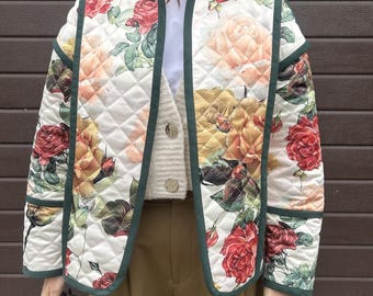 May include: Quilted jacket with a floral rose pattern in red, yellow, and peach hues on a white background. The jacket has a dark green trim and a diamond-stitched design. The jacket is open, revealing a white shirt and a beige cardigan.