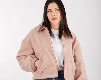 May include: A woman wearing a light pink fuzzy jacket with a zipper closure and a white t-shirt underneath. She is also wearing blue jeans.