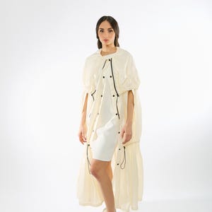 May include: A white long-sleeved dress with a white jacket over it. The jacket has a drawstring at the waist and a zipper closure. The model is wearing white sneakers.