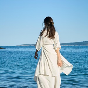 以下が含まれることがあります： 幅の広いゆったりとした袖とレイヤードスカートのデザインが特徴の、長く流れるような白いラップドレス。ウエストを結ぶタイプで、足首まで届く軽量で不透明な生地が、柔らかいシルエットを作り出しています。