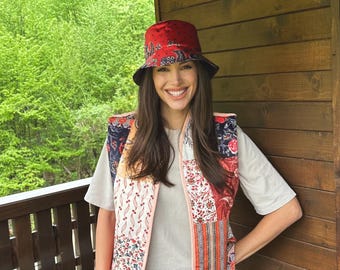 May include: A woman wearing a white t-shirt with a colorful patchwork vest and a red bucket hat. She is standing in front of a wooden wall.