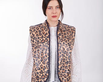 May include: A woman wearing a brown and black leopard print vest with a navy blue trim. She is also wearing a white blouse with eyelet detailing and blue jeans.
