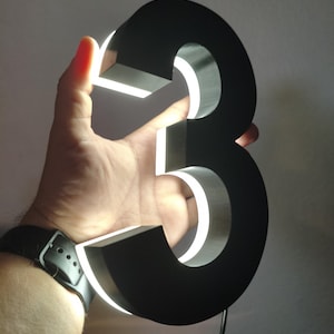 May include: A black, illuminated number three. The number is held up against a light background. The edges of the number are lit with a bright white light. The number is approximately 6 inches tall.