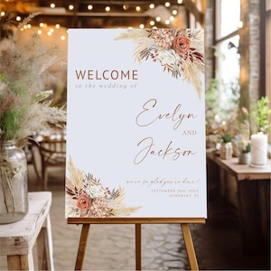 Könnte beinhalten: Ein Hochzeits-Willkommensschild auf einer Holzstaffelei. Das Schild ist hellblau mit Blumenarrangements in den Ecken. Der Text lautet "WELCOME to the wedding of Evelyn and Jackson" und "we're so glad you're here!" mit Datum und Ort.