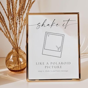 Könnte beinhalten: Ein gerahmtes Schild mit dem Schriftzug "shake it" in eleganter Schrift. Darunter eine Grafik im Polaroid-Stil mit zwei Herzen. Der Text lautet "LIKE A POLAROID PICTURE" mit der Anweisung "snap it, shake it and leave message".