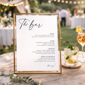 Könnte beinhalten: Ein gerahmtes Bar-Menü mit einem goldenen Metallrahmen. Das Menü listet Bier, Wein, Spirituosen und alkoholfreie Getränke auf. Der Text "The Bar" steht in Schreibschrift oben, mit dem Wort "cheers!" unten.