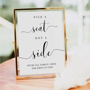 Könnte beinhalten: Ein goldgerahmtes Schild mit dem Text "PICK A seat NOT A side" in Schreibschrift. Der Text "WE'RE ALL FAMILY, ONCE THE KNOT IS TIED" ist ebenfalls enthalten. Geeignet für Hochzeiten oder besondere Anlässe.